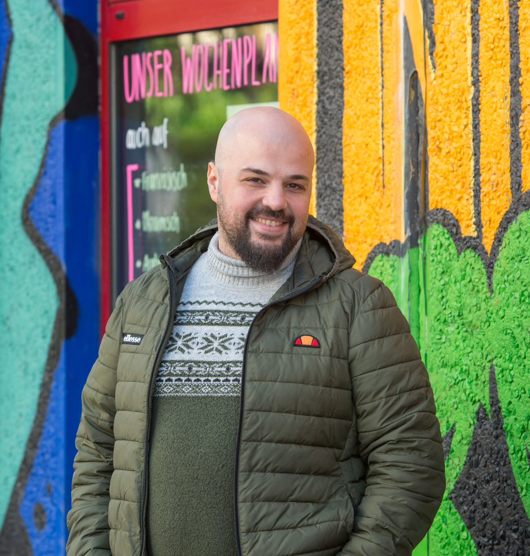 Hani Menzaljy blickt freundlich und aufmerksam in die Kamera. Er trägt eine olivegrüne Jacke und steht mit einer ruhigen und offenen Haltung vor einer bunten Wand mit Graffito.